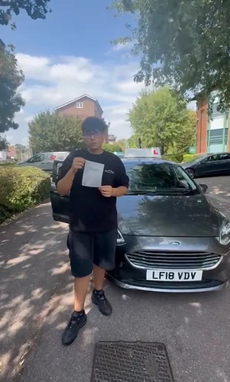 Young man in a black shirt holding his certificate next to a silver car in a parking lot